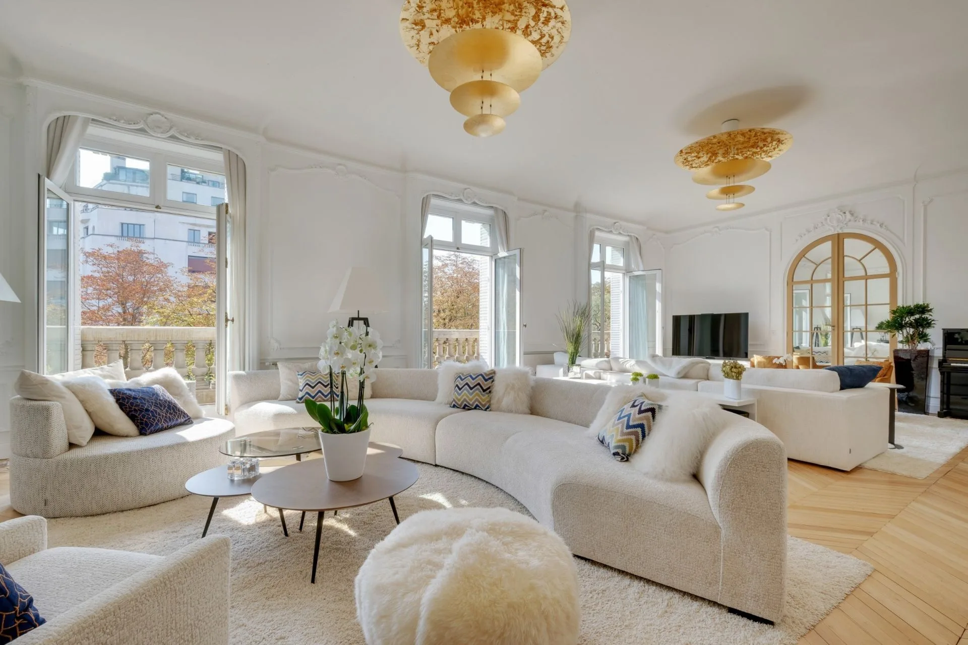 Grand salon haussmannien très lumineux - canapés blancs galbés, moulures, parquet en point de Hongrie et hautes fenêtres ouvrant sur un balcon